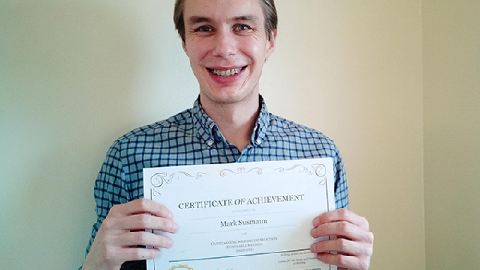 Mark Susmann, honorable mention for the 2020-2021 WAC Award for Outstanding Writing Instruction, holds his award certificate in front of a cream wall.